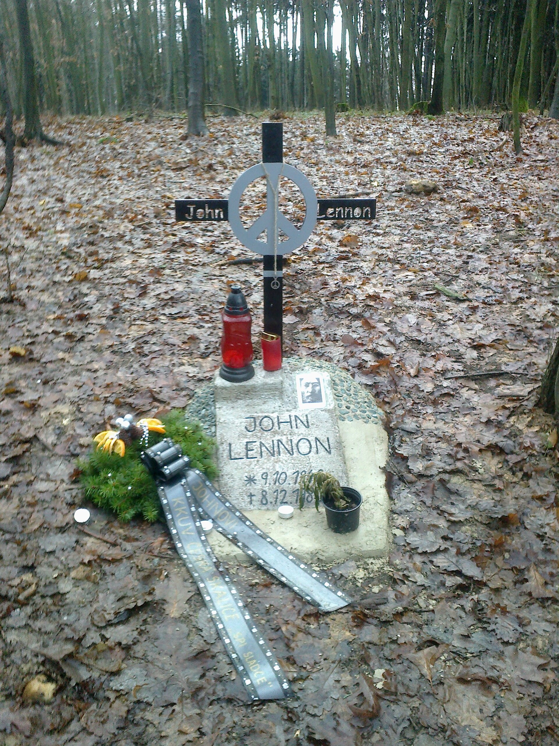 John Lennon memorial in the woods at Česká Skalice: a metal cross with a peace sign and a stone inscribed JOHN LENNON, with candles and a wreath