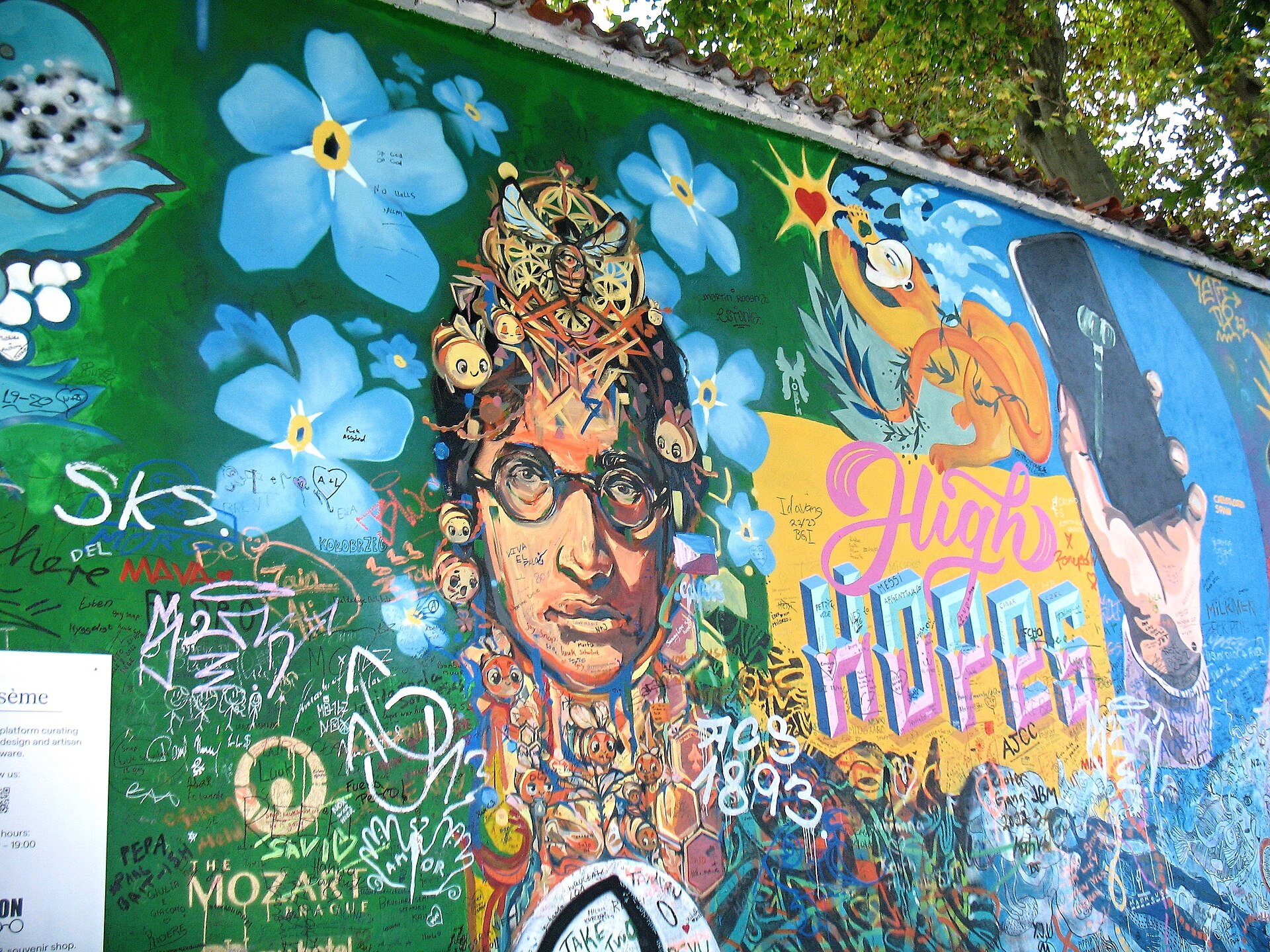 Painted portrait of John Lennon among blue flowers and visitors' writing on the Lennon Wall in Prague, October 2022