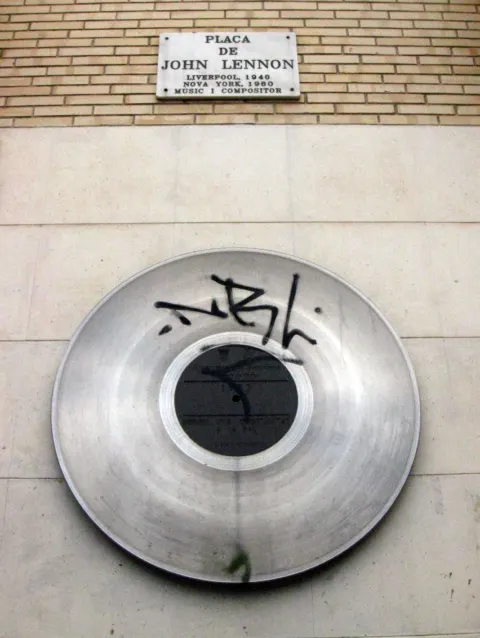 Record-shaped metal disc and marble street sign on a wall of Placa de John Lennon in Gracia, Barcelona